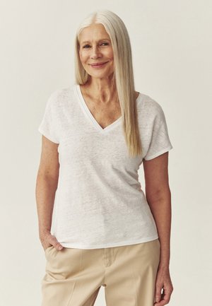 Femme âgée aux longs cheveux blancs porte un T-shirt blanc à col en V et un pantalon beige, souriant avec une main dans la poche sur un fond uni.