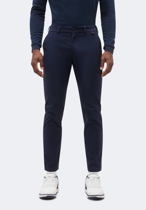 Bărbat purtând pantaloni bleumarin mulați, șosete albe și adidași albi, stând în fața unui fundal simplu și deschis la culoare.