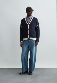 Cardigã azul-marinho com riscas vermelhas e bege, botões brancos, usado sobre uma camisa riscada, combinado com jeans azuis largos e sapatos pretos.