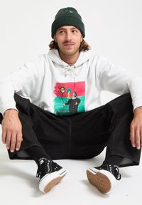 Hoodie gris avec un design illustré sur fond rouge et vert. Pantalons larges noirs, baskets noires avec des accents blancs et un bonnet vert.