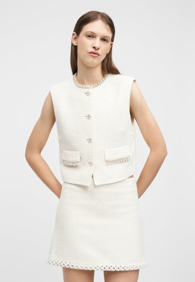 Femme portant un gilet blanc texturé sans manches avec des boutons en perle et une jupe assortie à ourlet perlé, debout les mains sur les hanches.