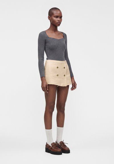 Femme debout portant un haut gris à manches longues, une mini-jupe beige taille haute boutonnée, des chaussettes blanches et des chaussures à plateforme marron sur fond blanc.