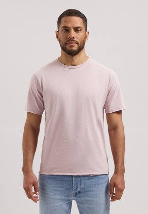 Man met kort haar en baard, gekleed in een lichtroze T-shirt met korte mouwen en lichtblauwe spijkerbroek, staand voor een effen achtergrond.