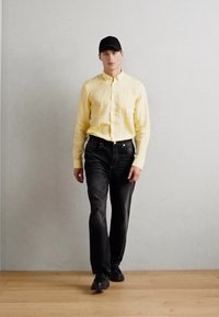 Yellow linen shirt, black denim jeans, black cap, and black shoes. Standing against a neutral background with wooden flooring.