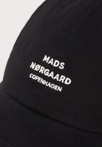 Melns beisbola cepures izgatavotas no kokvilnas ar baltu izšūtu tekstu: "MADS NØRGAARD COPENHAGEN." Iespējams, ar izliekto malu un teksturētu audumu.
