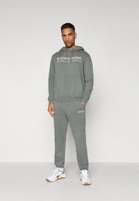 Homem em pé, vestindo um hoodie cinza da Björn Borg e calças de moletom, com ténis brancos, contra um fundo claro e simples.