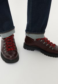 Mørkebrune lædervandre støvler med røde snørebånd, tykke sorte såler, båret med oprullede mørkeblå jeans mod en lys baggrund.