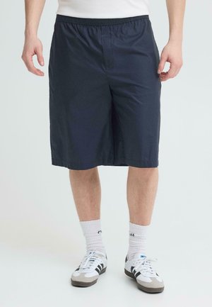 Mand iført marineblå knælange shorts, hvide crew-sokker og hvide sneakers med sorte striber, stående mod en ensfarvet baggrund.