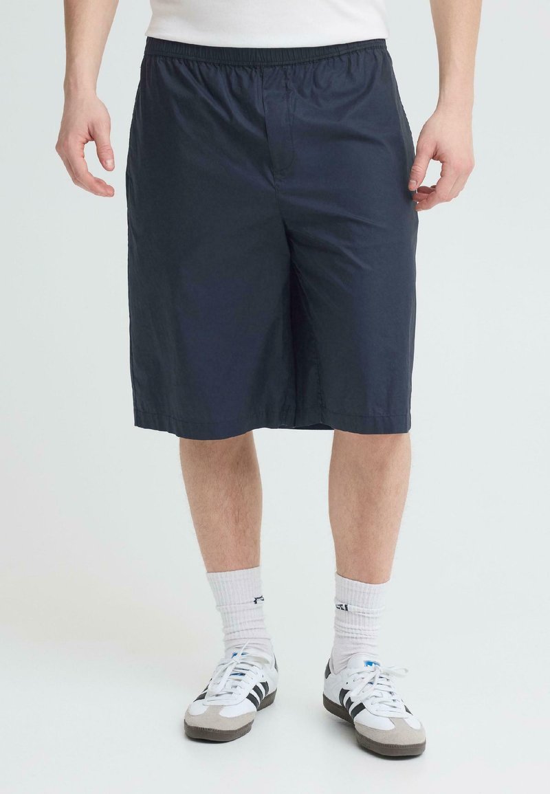 Homme portant un short bleu marine mi-cuisses, des chaussettes blanches montantes et des baskets blanches avec des bandes noires, debout devant un fond uni.