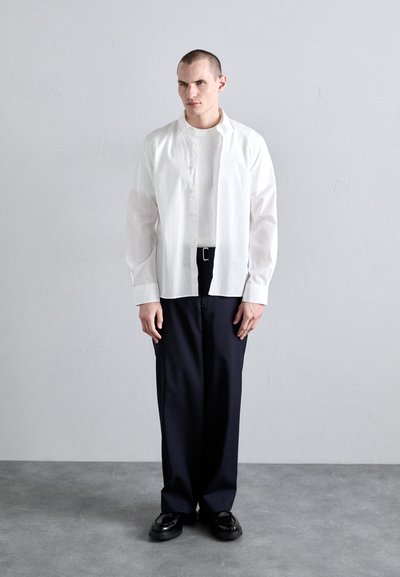 Camisa blanca de botones, con pliegues en los hombros, puesta sobre una camiseta blanca ajustada. Pantalones oscuros y holgados, zapatos negros, fondo neutral.