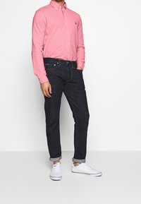 Muž v růžové košili s dlouhým rukávem a knoflíky, tmavých zvednutých džínách a bílých teniskách stojící proti jednobarevnému bílému pozadí.