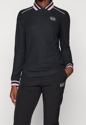 Personne portant un polo à manches longues noir avec un col et des poignets rayés rose et blanc, ainsi qu'un pantalon noir, tous deux avec les logos EA7 Emporio Armani.