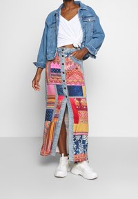 Kvinna som bär en patchwork-denimkjol med framknappar, en kortvit knuten topp, en överdimensionerad denimjacka och vita sneakers.