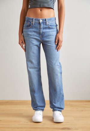 Persoon draagt lichtblauwe, rechtgesneden jeans met hoge taille, witte sneakers en een cropped gestreept topje tegen een effen achtergrond.