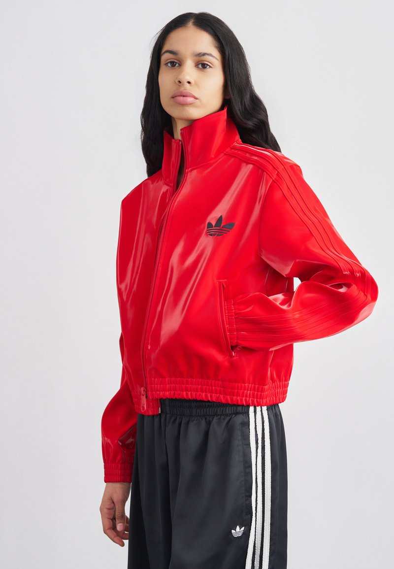 Rdeča sijoča jopa z zadrgo in visokim ovratnikom, z črnim logotipom Adidas in tremi kontrastnimi črtami na rokavih. Elastični manšeti in rob.