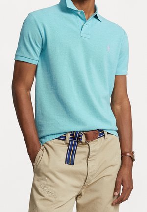 Homme portant un polo à manches courtes bleu clair, un pantalon beige usé avec une ceinture rayée bleue et jaune, et une montre en cuir marron au poignet droit.