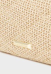 Vevd beige håndveske med en teksturert overflate, som har en gullhardware-logo plate. Designet er kompakt og rektangulært.
