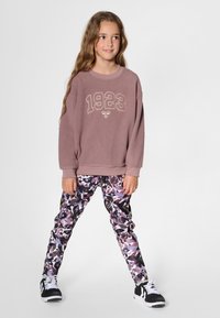Purple fleece sweatshirt with "1923" embroidered in gold, paired with multicolored camo leggings and black sneakers with white laces.