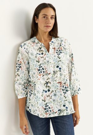 Femme portant un chemisier blanc à imprimé floral multicolore et manches trois-quarts, debout devant un mur clair uni.