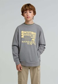 Felpa grigia con grafica gialla e design di teschio. Presenta un collo a giro e polsini a coste, abbinata a pantaloni cargo beige.