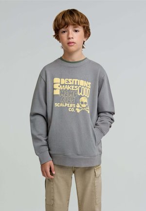 Sweatshirt gris avec texte graphique jaune et design de crâne. Dispose d'un col rond et de poignets côtelés, associé à un pantalon cargo beige.