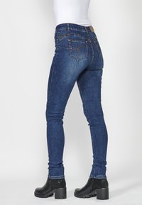 Skinny dark blue denim jeans with back pockets featuring a decorative stitch, paired with black ankle boots, displayed from the side.