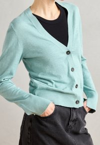 Mint green cardigan with a v-neck, five black buttons, and long sleeves; paired with black high-waisted pants. Soft texture and fitted design.