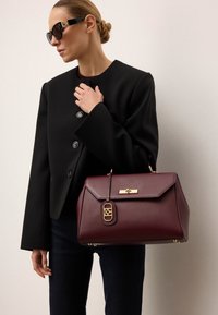 Burgundowa skórzana torebka o strukturalnym kształcie, z złotymi elementami i dekoracyjną metką. Noszona z czarną, oversize'ową kurtką i ciemnymi spodniami.