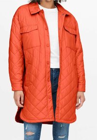 Cappotto arancione imbottito con colletto, dotato di due tasche sul petto, chiusura a pressione e spacco laterale, indossato sopra una maglietta bianca e jeans blu.