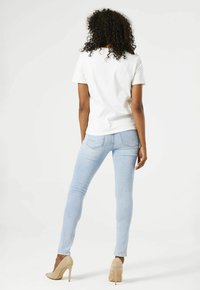 Camiseta de algodón blanca, jeans ajustados azul claro y zapatos de tacón beige. Cabello rizado visible en la parte posterior. Diseño simple y ajustado.