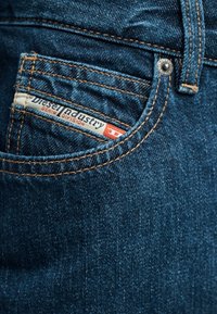 Κοντινό πλάνο στην τσέπη μπλε τζιν, με εμφανή την ετικέτα «Diesel Industry Denim Division» και πορτοκαλί ραφές.