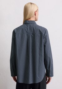 Kobieta z długimi blond włosami, ubrana w czarną koszulę z guzikami w mały niebiesko-biały geometryczny wzór, pokazana od tyłu.