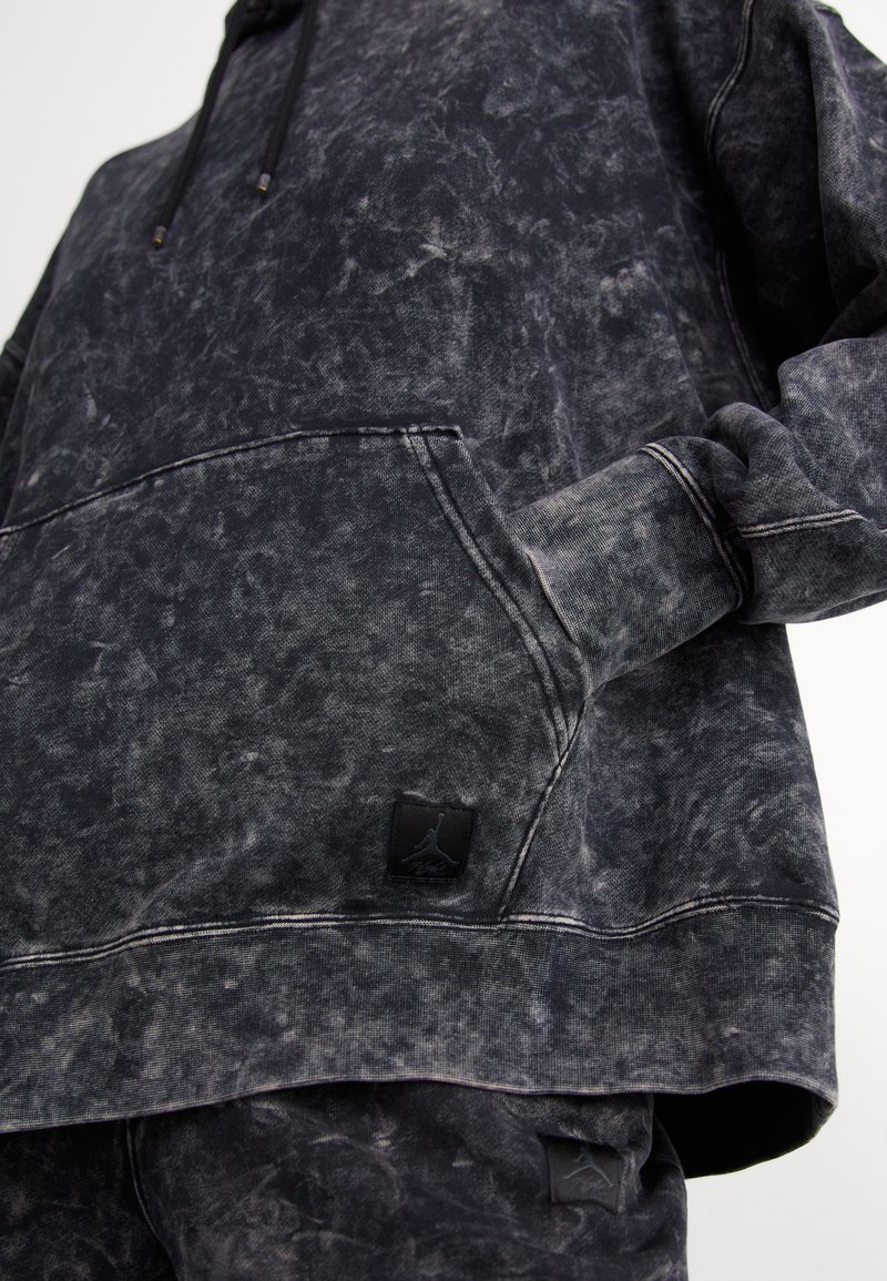 Sudadera negra con efecto stonewashed, con bolsillo canguro, puños acanalados y parche negro con logo. La tela tiene un aspecto suave y texturizado.