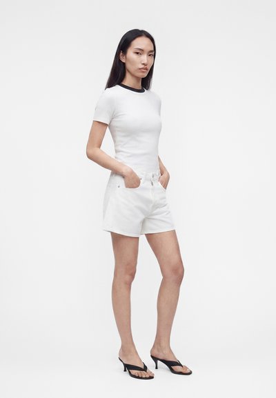 Γυναίκα που στέκεται με λευκά σορτς και λευκό μπλουζάκι με μαύρο γιακά, χέρια στις τσέπες, φοράει μαύρα πέδιλα με τακούνι, με απλό φόντο.