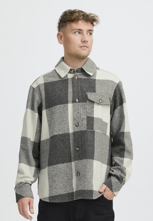 Ung mand iført en sort, hvid og grå storternede skjorte med knapper og en brystlomme, der kigger mod højre med hånden i lommen.