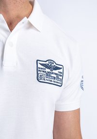 Bílá polo košile vyrobená z texturované látky. Obsahuje modré vyšívané nášivky na hrudi, které zobrazují branding a zeměpisné souřadnice.