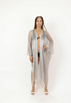 Vrouw met lange vlechten staat tegen een witte achtergrond, draagt een zwarte bikini en een zilveren, netachtige, met franjes afgewerkte cover-up met open voorkant.