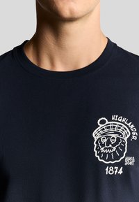 Marineblå bomulds T-shirt med et hvidt grafisk motiv af et skægget ansigt i hat, med teksten "HIGHLANDER" og "1874". Rund halsudskæring og ribbet tekstur.