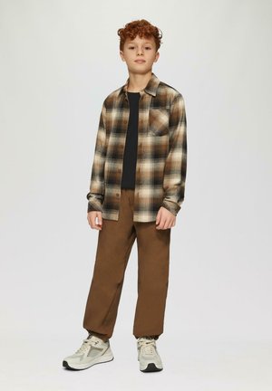 Geruit overhemd in tan- en bruintinten, zwart T-shirt eronder, bruine broek en beige sneakers met textuuraccenten. Gathered cuffs op de broek.