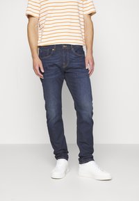 Jeans de mezclilla azul oscuro con corte ajustado, con un desvanecimiento sutil. Combinados con una camiseta de rayas de manga corta y zapatillas blancas.