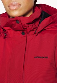 Chaqueta impermeable roja con un alto cuello, forro negro, botones a presión y un logo bordado de "Didriksons" en el pecho.