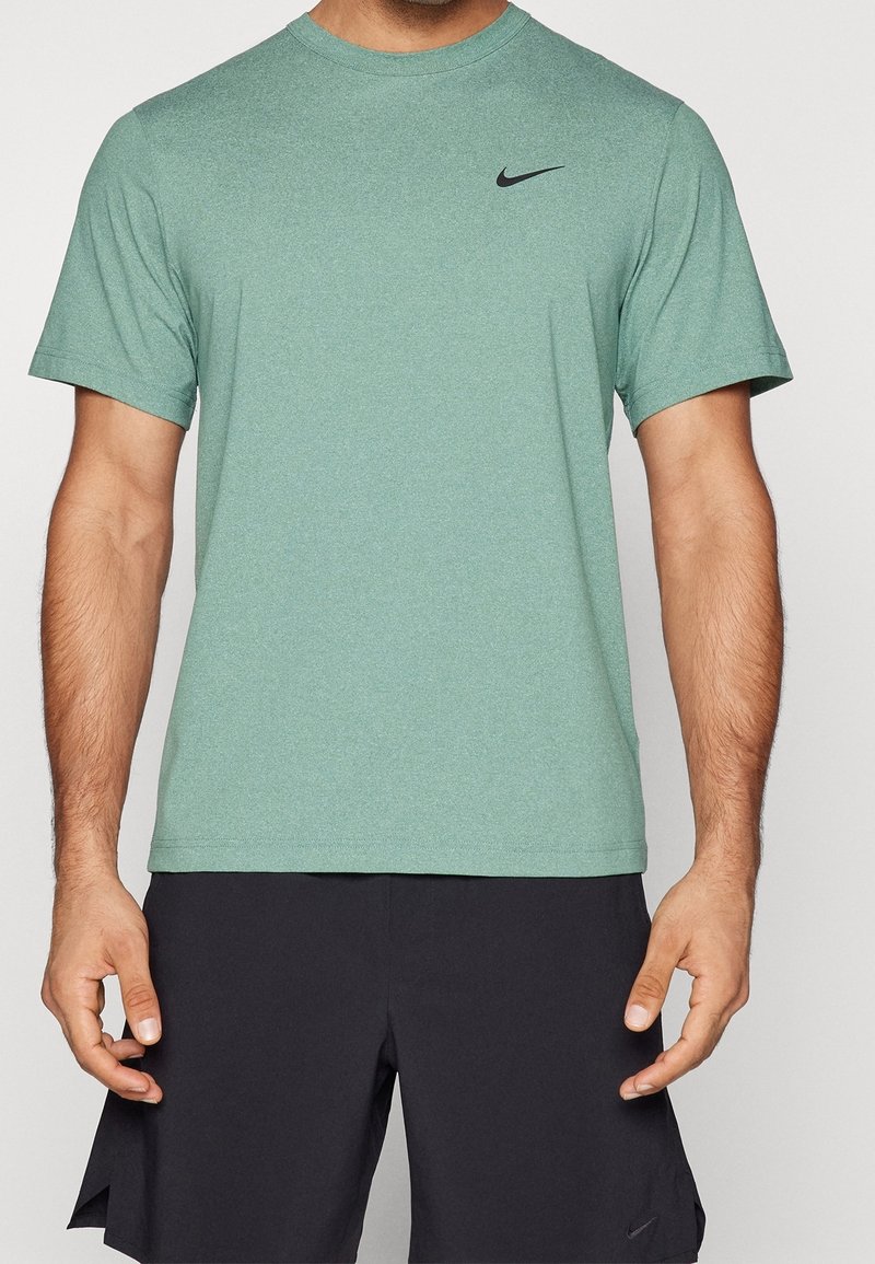 Homme portant un t-shirt de sport à manches courtes vert de Nike et un short noir de Nike, debout avec les bras détendus le long du corps.