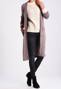 Un long cardigan ouvert à l'avant en tricot texturé gris, avec deux poches, superposé sur un pull en rib crème et un jean skinny noir, associé à des bottines noires.