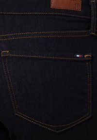 Jeans de mezclilla azul oscuro con costuras amarillas, que presentan una etiqueta de marca de cuero y un pequeño logotipo multicolor en el bolsillo trasero.