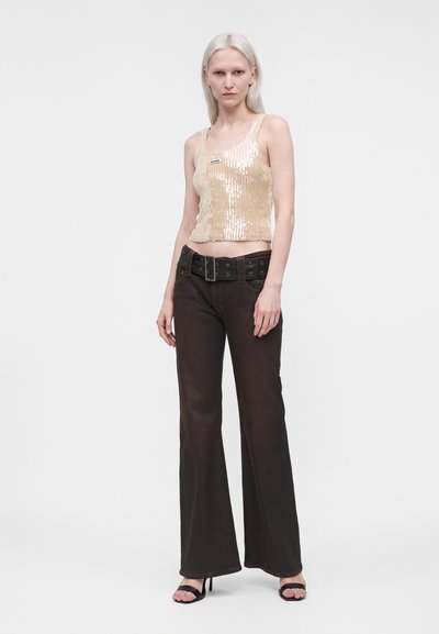 Femme aux cheveux clairs portant un haut sans manches beige à sequins, une large ceinture noire, un pantalon flare foncé et des talons ouverts noirs, debout sur fond blanc.