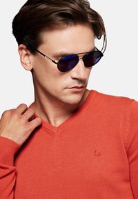 Czarne okulary przeciwsłoneczne w stylu aviator z niebieskimi soczewkami i srebrnymi akcentami, noszone przez osobę w jasnopomarańczowym swetrze V-neck z dzianiny.