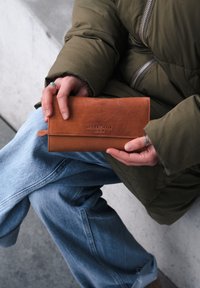 Braune Lederbrieftasche mit geprägten Buchstaben, rechteckige Form, glatte Oberfläche, gehalten in einer Hand mit silbernen Ringen, im Hintergrund eine grüne Steppjacke sichtbar.