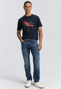 Camiseta de algodón azul marino con texto gráfico en blanco y rojo, combinada con jeans de mezclilla azul claro y zapatillas blancas. Un conjunto simple y casual.