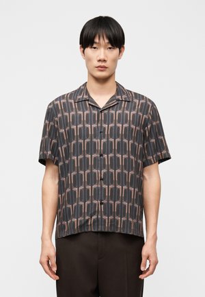 Jeune homme portant une chemise à manches courtes, boutonnée, avec un motif géométrique dans des tons de gris et de marron sur un fond uni.