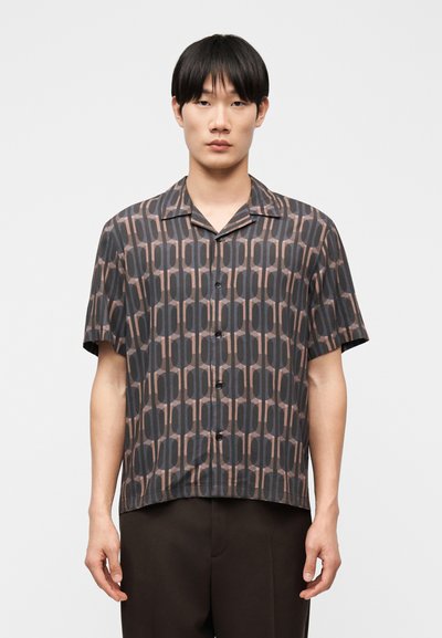 Jeune homme portant une chemise à manches courtes, boutonnée, avec un motif géométrique dans des tons de gris et de marron sur un fond uni.